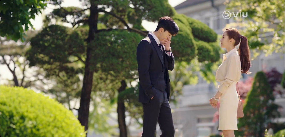 Man in suit and woman in beige dress stand in a lush garden with trimmed trees and building in background, sunlit and contemplative mood.