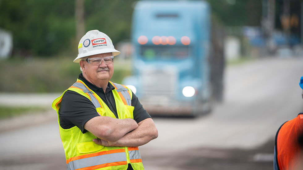 Construction worker in safety gear stands with arms crossed on a road, with a large blue truck approaching in the background.