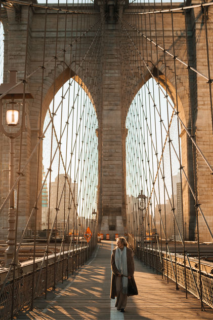 Wide-angle sunrise shot of the Brooklyn Bridge walkway with its iconic stone arches and a woman in the distance, NYC
