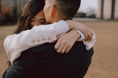 Close-up of couple embracing at the DUMBO waterfront, bride's diamond engagement ring and gold bracelet visible, NYC