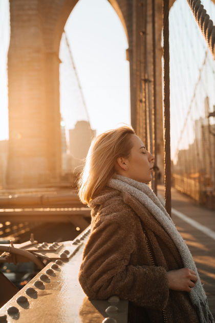 Portrait of a woman in a teddy coat standing on the Brooklyn Bridge at sunset with the Manhattan skyline, NYC