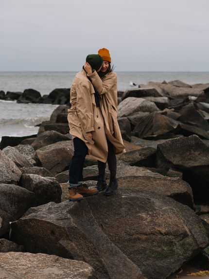 Couple standing on large rocks by the ocean at Coney Island in winter, grey sky and ocean behind them, NYC couple shoot