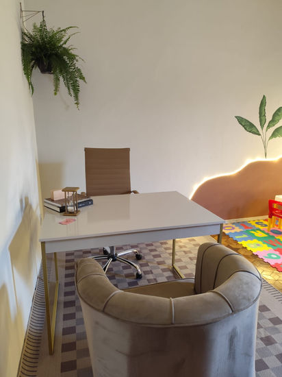 foto de uma sala que aparece uma cadeira de escritorio no fundo, na frente uma mesa e na frente uma poltrona. Uma samambaia ao fundo