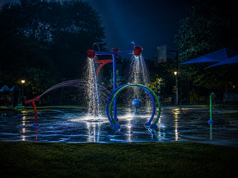 An empty splash pad at Janssen Park glows beneath a moonlit twilight sky as running water reflects across the wet pavement.
