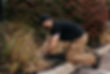 Employee black cap kneeling, tending to garden bed to freshen mulch along curb.