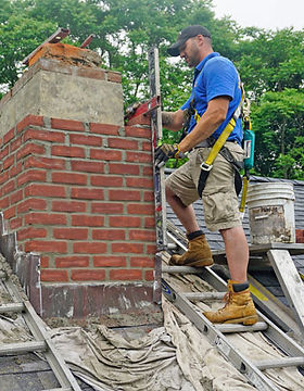 Mason in a safety harness on a roof ladde rebuilding a red brick cimney above the roofline