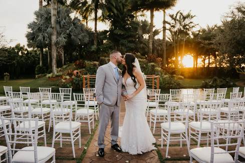 Bride and groom share a kiss at their sunset wedding ceremony at Longan's Place in Miami, surrounded by lush greenery and white chairs, captured by Aleks Victoria Photography.