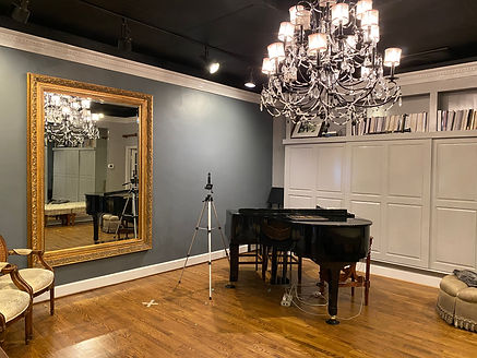 A picture of the Amy Murphy Studio! Featuring a grand piano, a bookcase spanning the entire side wall, hard wood floors, and an enormous mirror. There is also filming equipment if a singer needs to self tape