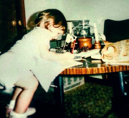 Emily Goff as a 2-year old child with brown hair, a white outfit, and pink shoes, coloring at a table with a smile on her face as her orange cat watches on. It's a very 1980s looking room.