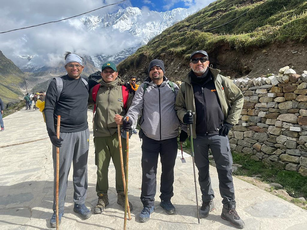 Four men with hiking gear and walking sticks smile on a stone path. Snowy mountains, grassy hills, and partly cloudy sky in the background.