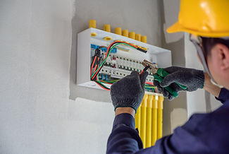 A worker in a hard hat and gloves using pliers to work on a circuit breaker panel mounted on a wall, with yellow conduit pipes running alongside.