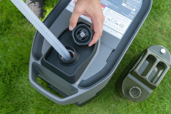 Hand inserts a nozzle into a gray container on green grass. Another closed container is nearby. Labels with text are visible on the container.