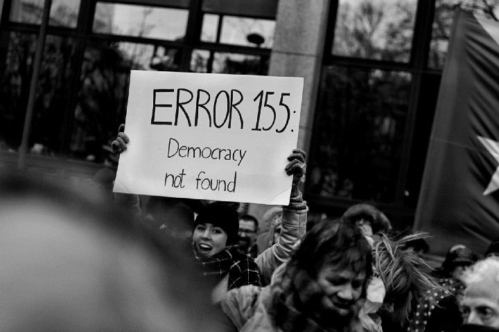 a woman in a beanie and scarf, in a crowd of people, holding up a sign that reads ERROR 155: Democracy not found