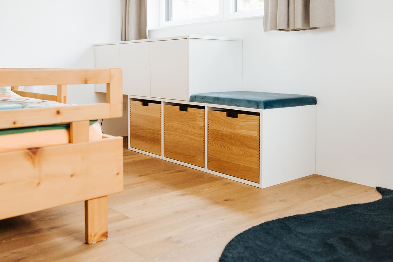 Kinderzimmer mit funktionalem, freundlichem Mobiliar in Naturtönen. Links im Bild steht ein Bettgestell aus hellem Holz mit grüner und orangefarbener Bettwäsche. Rechts davon eine clevere Sitzbank mit integrierten, herausziehbaren Holzkisten zur Aufbewahrung. Die Sitzfläche ist gepolstert und mit einem dunkelgrünen Kissen belegt. Dahinter befindet sich ein weißes Sideboard unter dem Fenster, das zusätzlichen Stauraum bietet. Der Raum hat einen warmen Holzfußboden und helle Vorhänge, die das Fenster seitlich einrahmen.