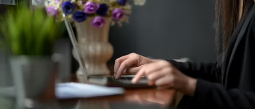 Hands typing on a laptop in a dimly lit room. Vase with purple and pink flowers in the background. Green plant and papers nearby.