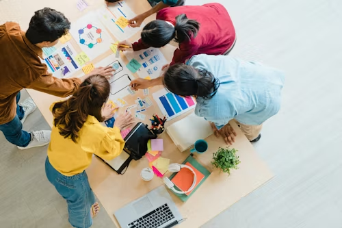 Five people collaborate on a project at a table with charts and sticky notes. Bright clothes, laptop, and plant in view, suggesting creativity.