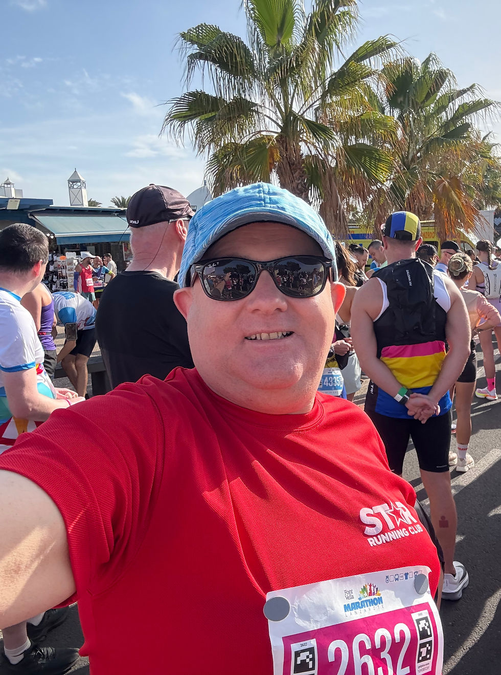 Paul wearing a red Star Running Club t-shirt, RayBan sunglasses and a blue cap. His race number is affixed to his t-shirt and there's a palm tree and other runners behind him.