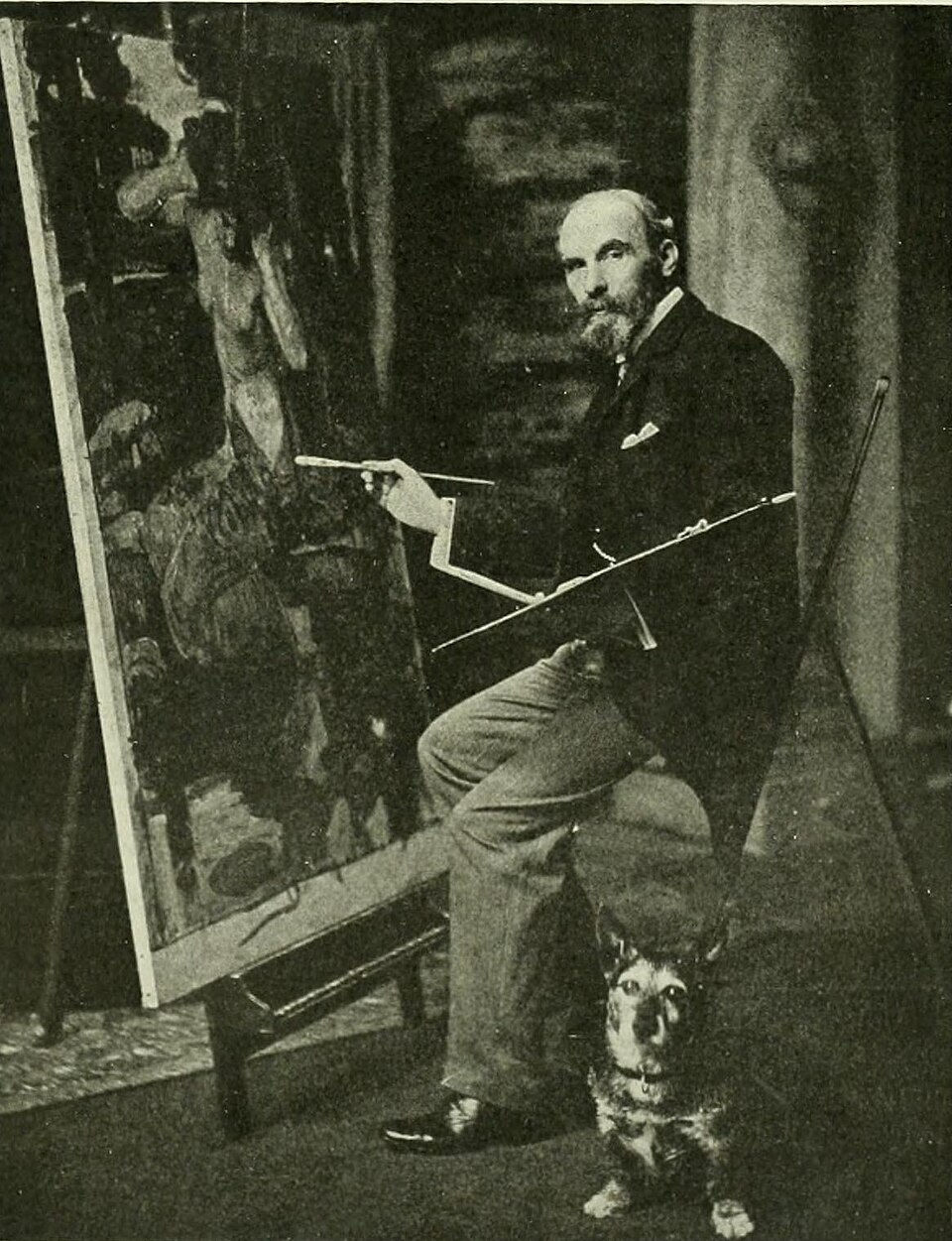Bearded painter seated at an easel in a dim studio, brush in hand, with a small dog at his feet in a black-and-white photo