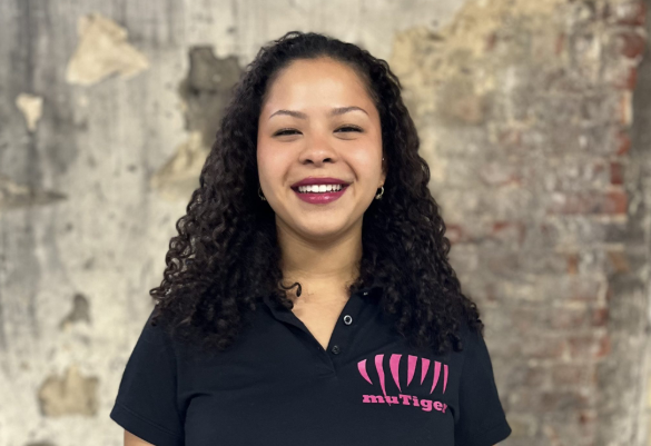 Eine junge Frau mit lockigen dunklen Haaren steht lächelnd vor einer Wand mit Putz- und Backsteinstruktur. Sie trägt ein schwarzes Polo-Shirt mit pinkem muTiger-Logo