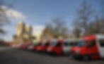 Mehrere rote Bürgerbusse stehen auf einem Platz vor dem historischen Dom in Münster in einer Reihe nebeneinander. Die Fahrzeuge sind unterschiedlich gestaltet und tragen Logos sowie Beschriftungen verschiedener Bürgerbusvereine. Im Hintergrund sind der Dom, Bäume ohne Laub und ein blauer Himmel zu sehen.