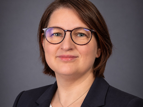 Porträt von Norma mit kurzen braunen Haaren und Brille vor einem dunkelgrauen Hintergrund. Sie trägt einen dunkelblauen Blazer und eine weiße Bluse.