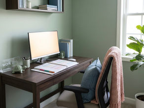 Home office with a desk, monitor, books, and a "work only" sign. A chair with a pink blanket, green plant, and soft natural light. Cozy mood.