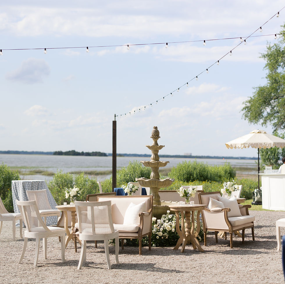 Elegant outdoor seating with white chairs, sofas, and a fountain centerpiece. String lights above. Lake and greenery in the background. Calm mood.