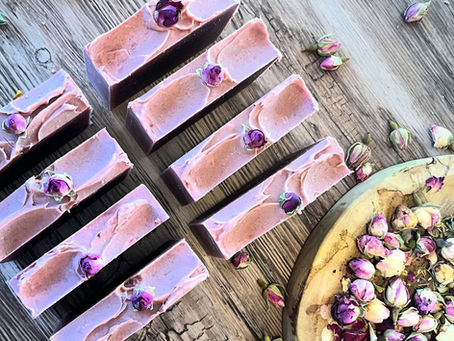 pink bars of soaps laid out on a rustic brown wooden table with rose buds on them a plate of rose buds by its side