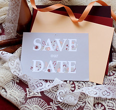 Eine Save the Date-Karte zur Hochzeit in Grau, Apricot und Rot liegt auf weißem Spitzenstoff vor farbigen Umschlägen