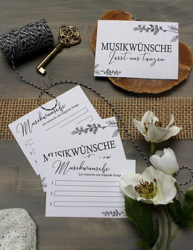 Moderne Musikwunschkarten in Schwarz und Weiß liegen auf grauem Untergrund neben weißen Blumen und einem goldenen Schlüssel