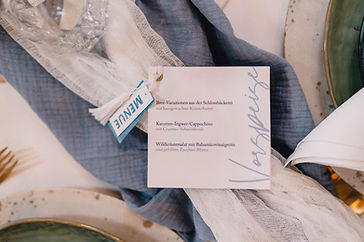 Eine elegante Menükarte in Blau und Weiß im Fächerformat liegt auf einem blauen Tuch auf einem Hochzeitstisch