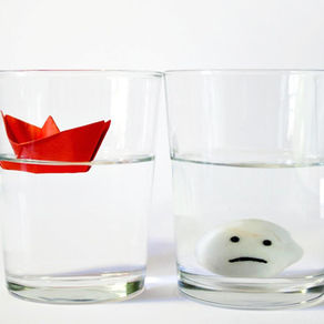 Two glasses of water side by side, one with a floating red paper boat and one with a sinking sad face object, symbolizing optimism and pessimism