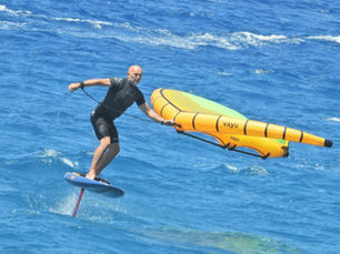Wing-foiler gliding above the sea on a hydrofoil board, holding a bright yellow inflatable wing, with another rider in the background.