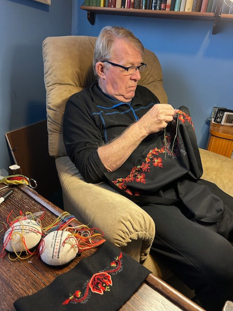 An older man sits in an armchair, hand-embroidering a bunad piece with red floral patterns, with colorful embroidery threads and tools on a table beside him.