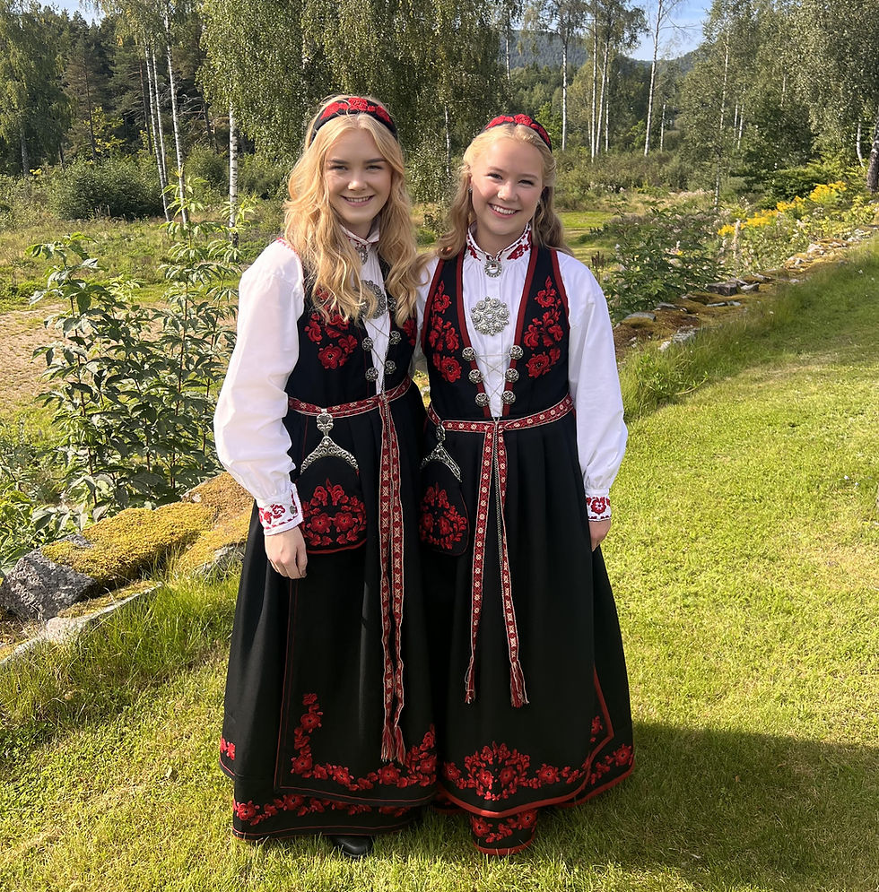Two smiling girls in traditional Raudsau bunads with red embroidery stand on a sunny lawn, with birch trees and mountains in the background
