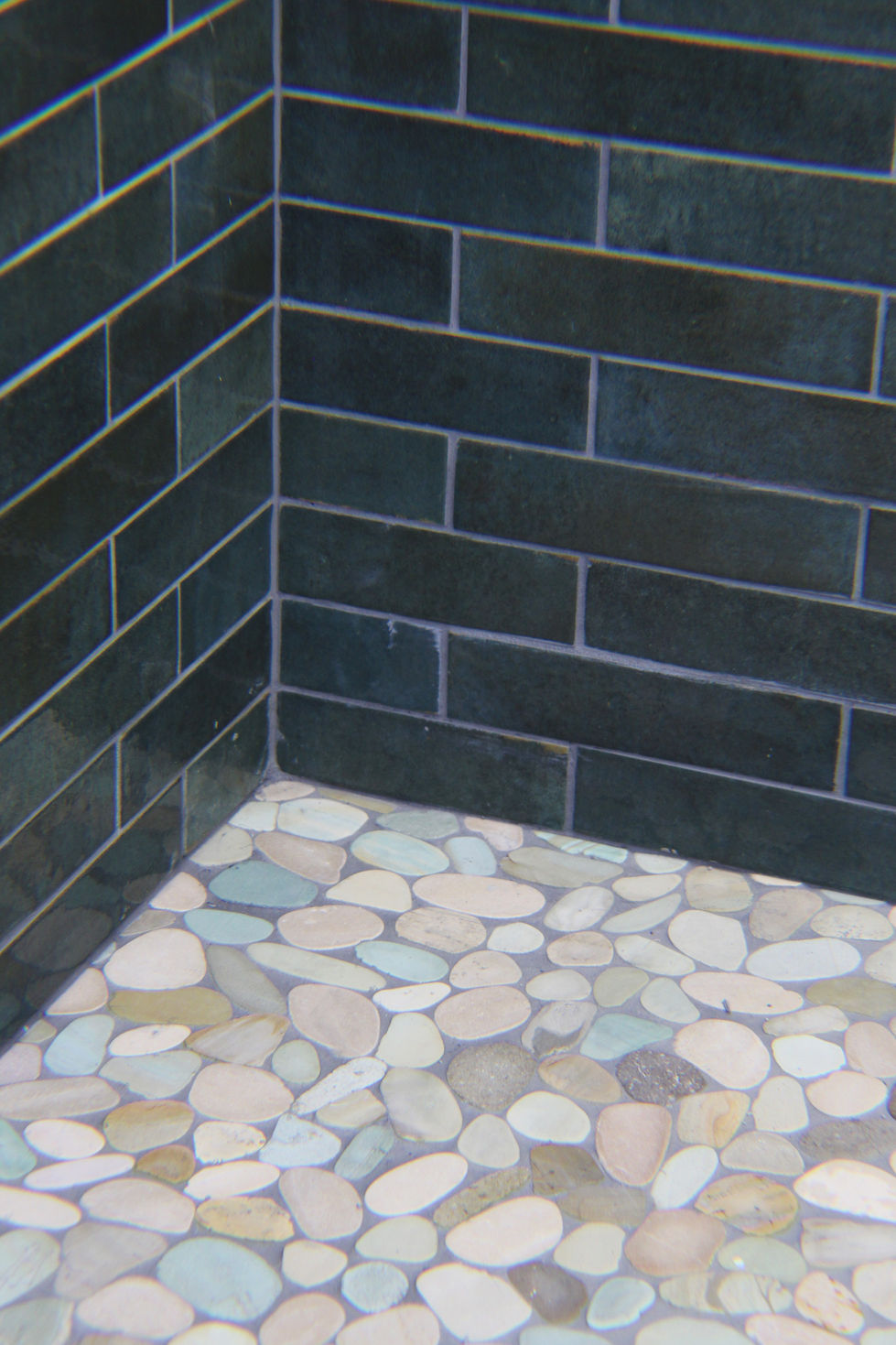 Close up of walk-in shower corner where the green 2x8 wall tiles meets the pebble mosaic floor tiles