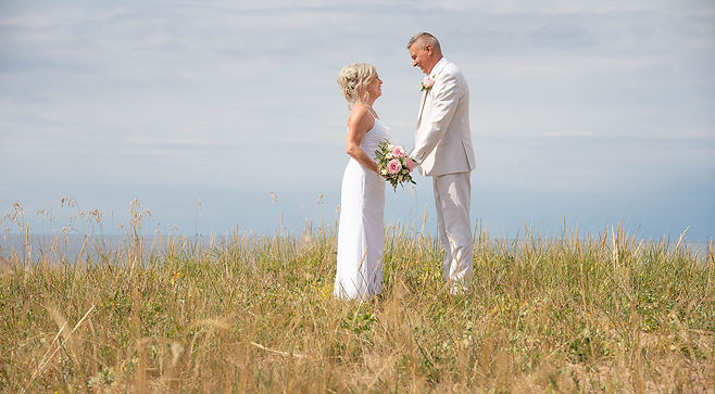 Un couple de mariés vêtus de blanc se fait face et se tient par les mains dans un champ herbeux, sous un ciel clair.