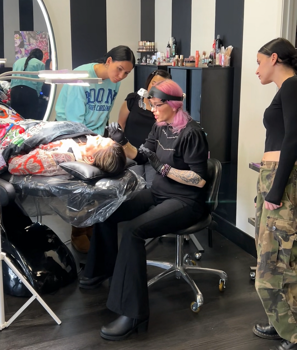 Pink-haired artist applies eyeliner to a client reclining on a salon chair, with two observers in a striped studio.