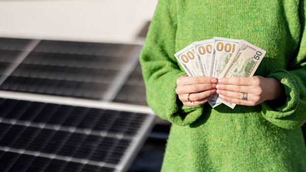 A woman standing next to solar panels holding onto a handful of one-hundred dollar bills.