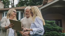 An insurance inspector shows a clipboard to a couple in front of their large home.