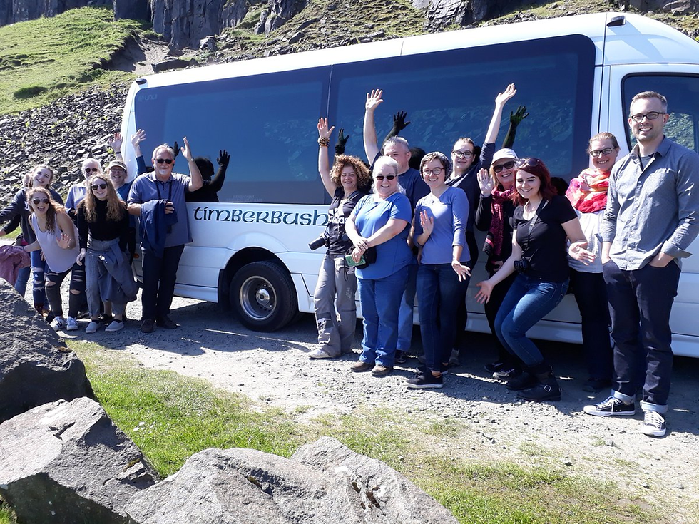 A group of people cheerfully pose by a white Timberbush tour bus. They wave in a rocky, grassy setting under a clear blue sky.