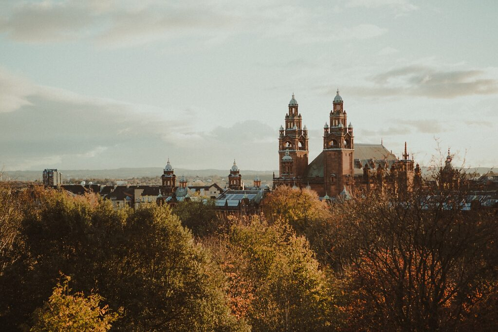 Leafy foreground with red-brick cathedral towers in the background under a cloudy sky; autumn hues create a serene, historic ambiance.