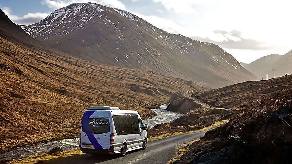 A white van travels a winding road through a mountainous landscape, with a river beside it. Snow caps the peaks under a cloudy sky.