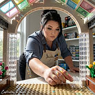 An adult designer presses the final transparent LEGO piece into a glass-roofed botanical conservatory as morning light passes through the model.