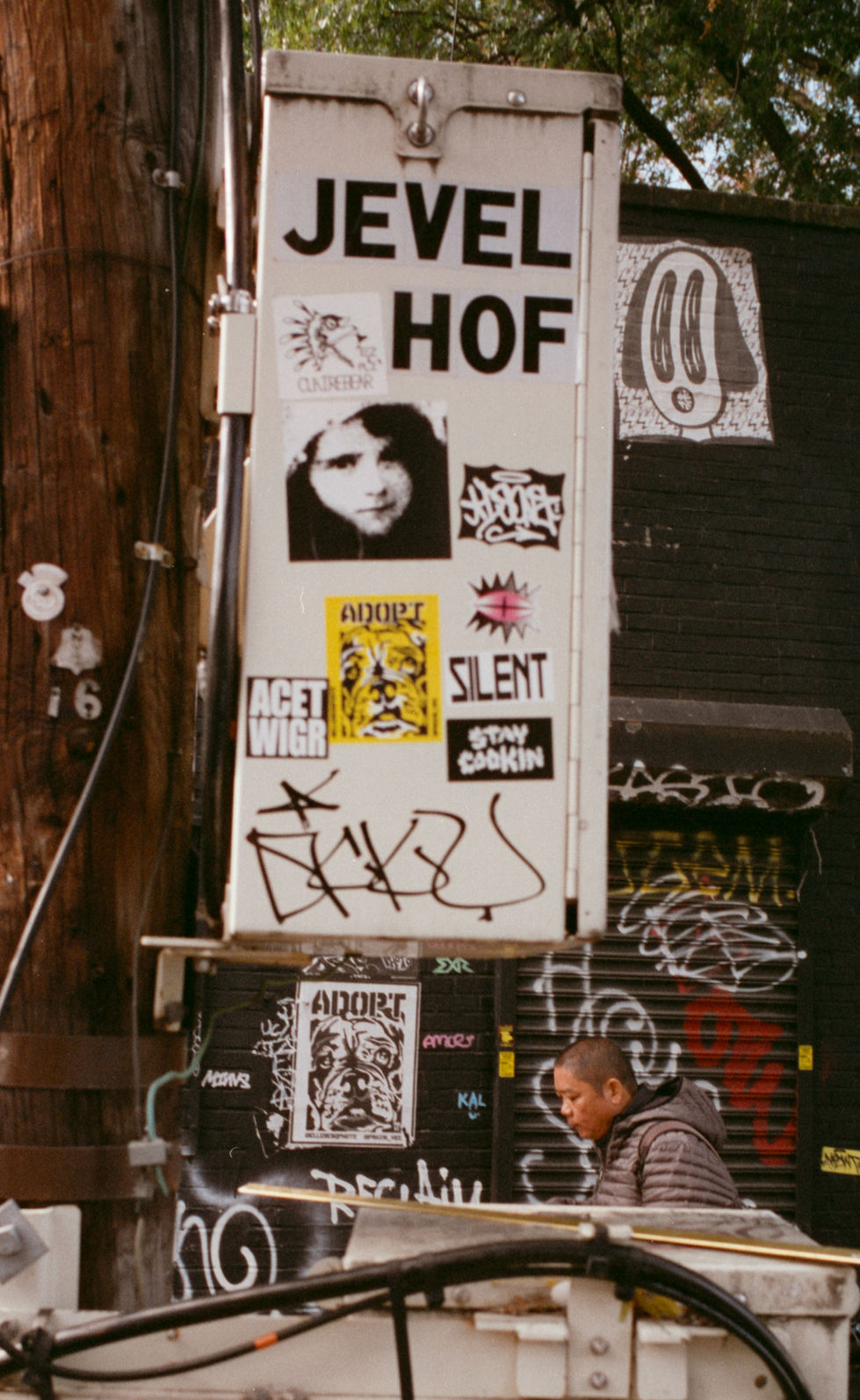 Person DJing beneath "JEVEL HOF" "SILENT" sign, covered in street art.