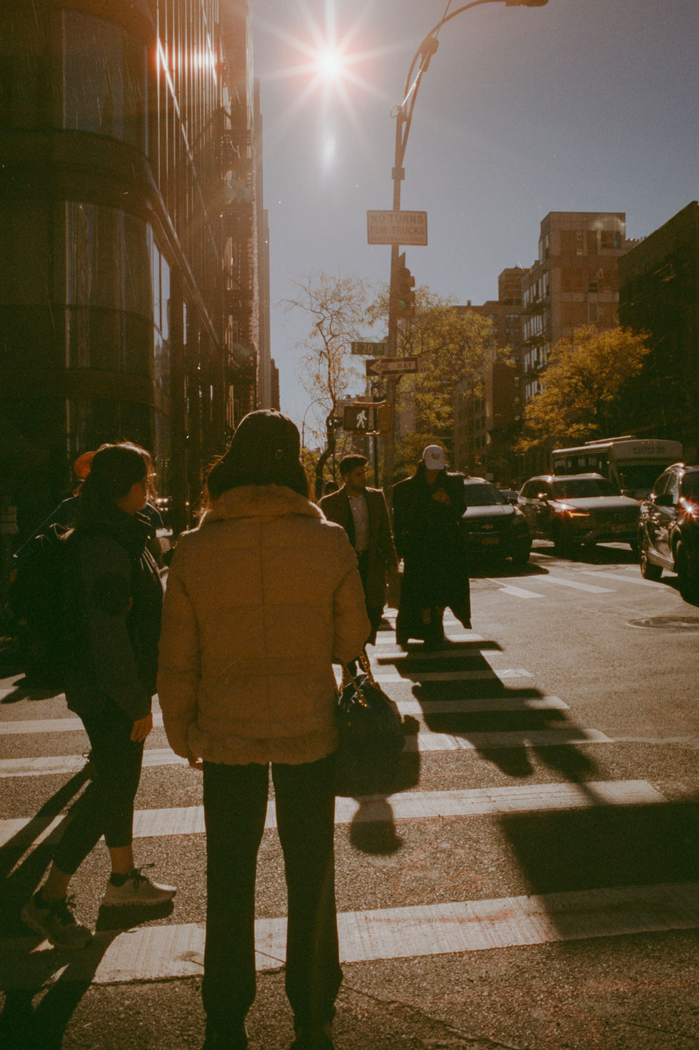 People crossing sunlit city street, casting long shadows, with buildings.