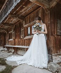 Braut in weißem Hochzeitskleid steht vor Holzgebäude, hält Blumenstrauß in der Hand und schaut nach unten