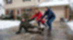 Two people rush to help an older man slipping on an icy driveway outside a snowy house, his face tense with pain