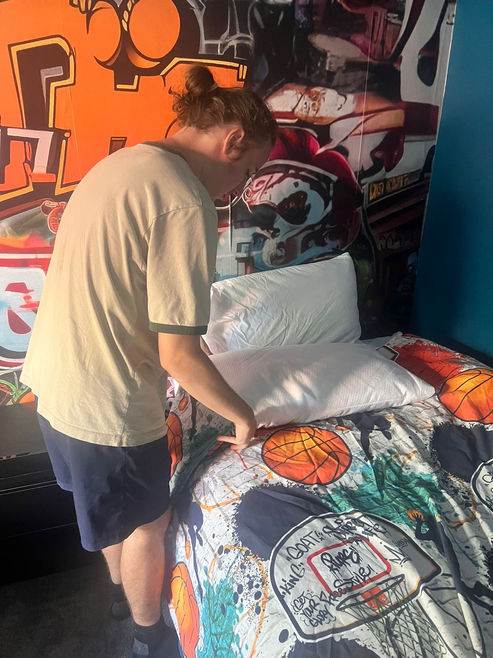 A person in a tan shirt and shorts making a bed with a colorful patterned bedspread and white pillows in a room with graffiti wall art.