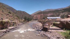 Stone bridge crossing a river through a mountain valley, with rolling hills, scattered buildings, and a clear blue sky in the background.